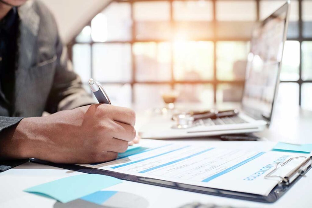 A person sitting at a kitchen table reviewing insurance paperwork and a laptop, with family members in the background.